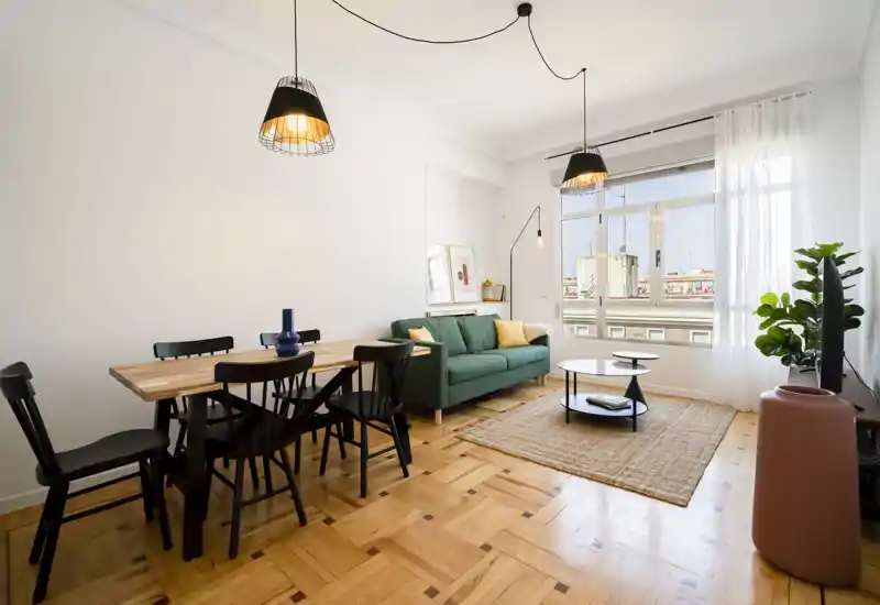 Salón luminoso y con estilo, con sofá verde, luz natural por una gran ventana, alfombra de yute y mesa de comedor; perfecto para relajarse y recibir invitados.