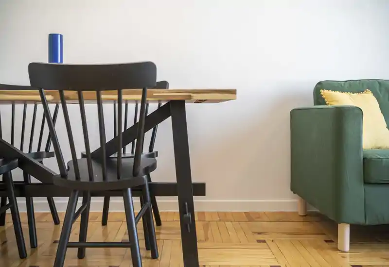 Zona de estar luminosa y minimalista con mesa de madera, sillas negras y un sofá verde con cojín amarillo — estilo moderno y limpio ideal para anuncios de alquiler.