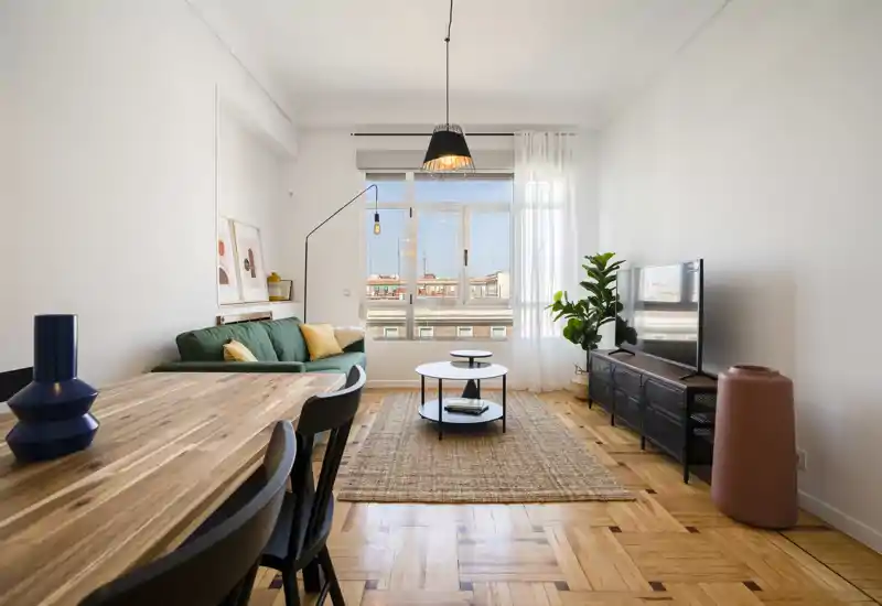 Sala de estar moderna y luminosa con sofá verde, amplia ventana con luz natural, alfombra de fibras y mueble de TV — estilosa y acogedora.