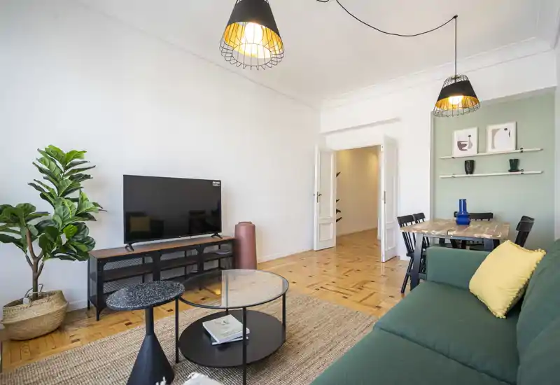 Sala de estar luminosa y con estilo con sofá verde, mueble de TV, mesas de centro y mesa de comedor al fondo; lámparas colgantes cálidas y plantas crean un ambiente acogedor.