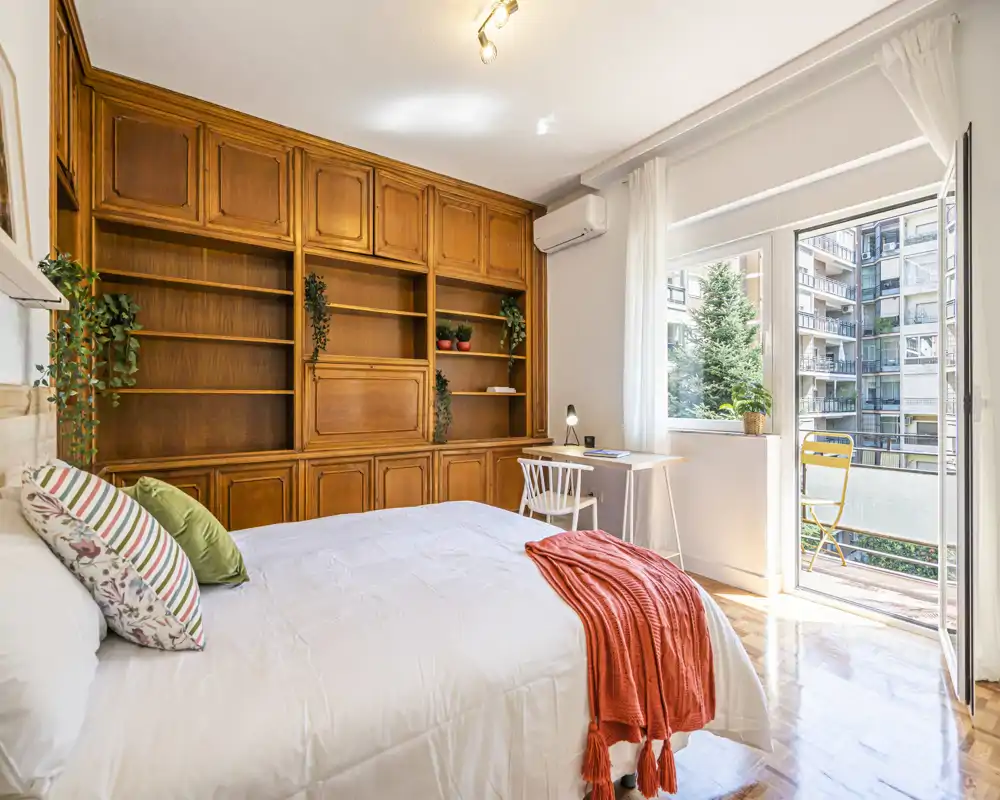 Bright, stylish bedroom with a large built-in wooden shelving unit, a neatly made bed with colorful cushions and a coral throw, plus a small desk by the window opening onto a sunny balcony.