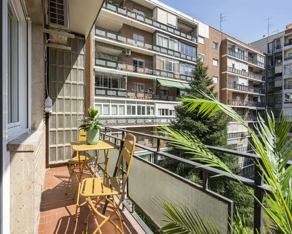 Sunny balcony with a small table and two chairs, potted plants and a railing overlooking neighboring apartments — ideal for morning coffee.