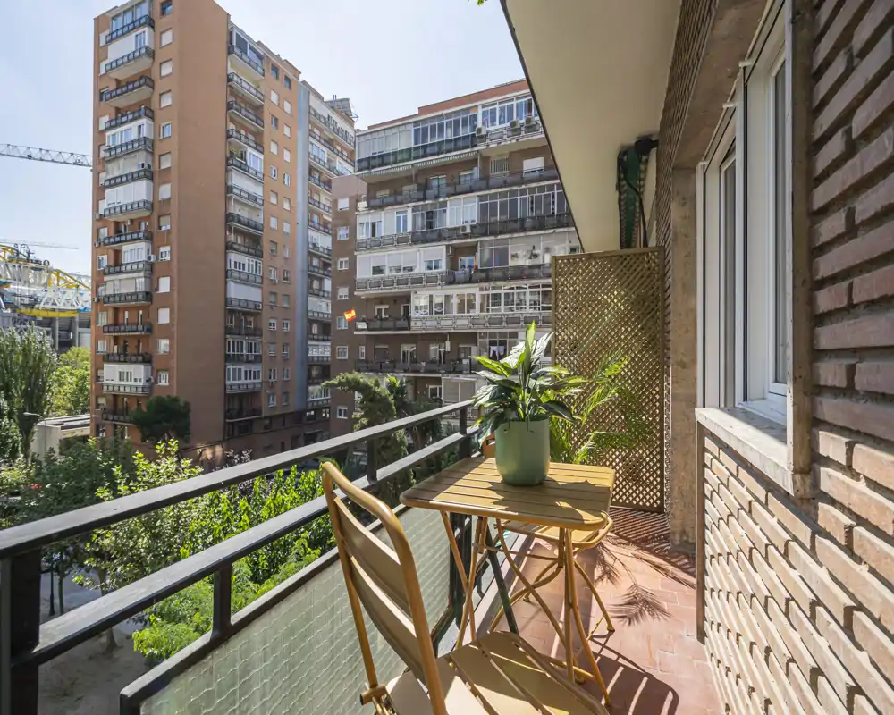Sunny city balcony with a small wooden table and two chairs, potted plants, and a view over neighboring apartment buildings — ideal for morning coffee.