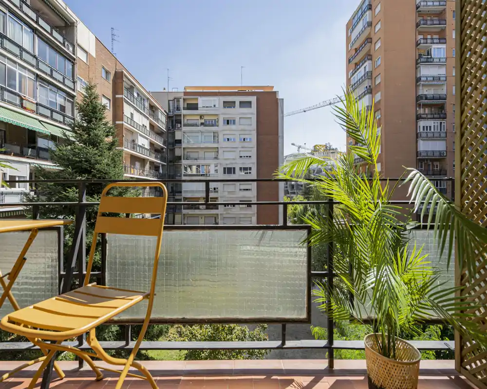 Sunny city balcony with a small yellow bistro set and potted palm, overlooking neighbouring apartment buildings — ideal for morning coffee.