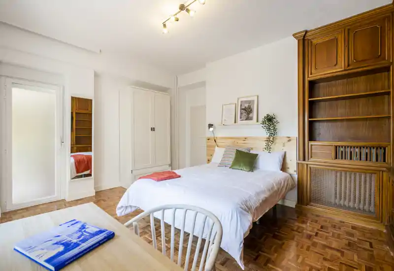Bright, styled bedroom with a double bed, wooden headboard and large built-in shelving. Natural light and warm parquet flooring create a welcoming, well-appointed sleeping space.