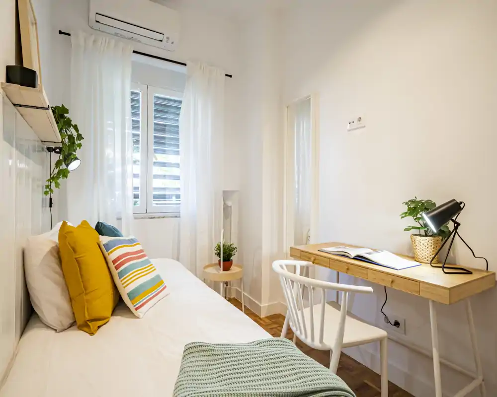 Bright, neatly staged bedroom with a single bed, colorful cushions and a small wooden desk by the wall — ideal for a student or short-stay guest.