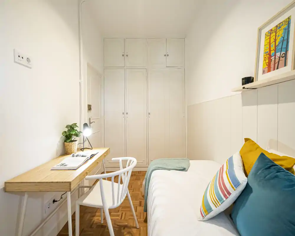 Bright, compact bedroom with a neatly made single bed, colorful cushions and a small wooden desk by the wall — ideal for a student or short-stay guest.