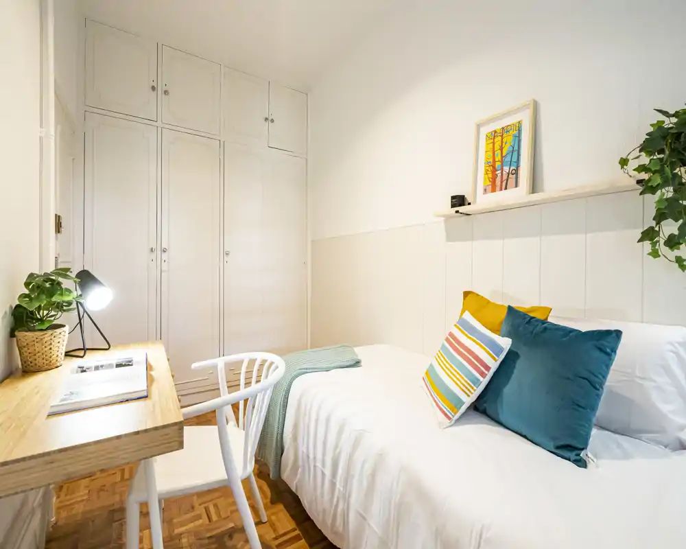 Bright, styled single bedroom with a comfortable bed, colorful cushions and a small wooden desk by the window — ideal for a student or solo traveler.