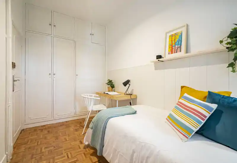 Bright, neatly styled single bedroom with built-in wardrobes, a compact desk, and colorful cushions and textiles creating a welcoming atmosphere.