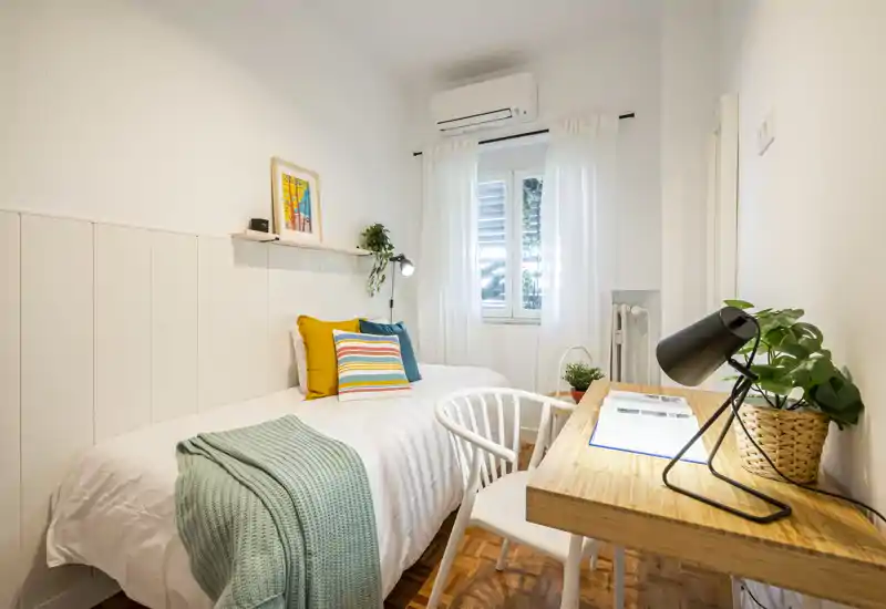 Sunny, well-styled single bedroom with a comfortable bed, colorful cushions and a wooden desk by the window — ideal for work and rest.