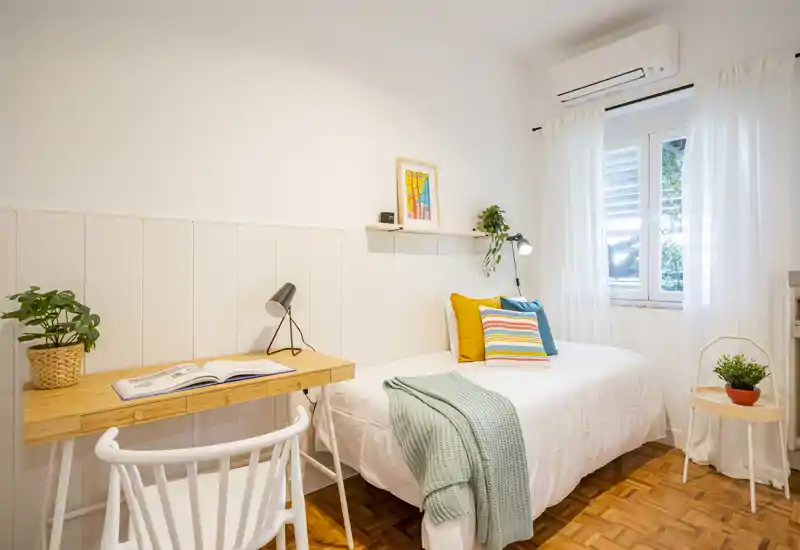 A bright, neatly styled single bedroom with a comfortable bed, colorful cushions and a small work desk by the window — ideal for a student or solo traveler.