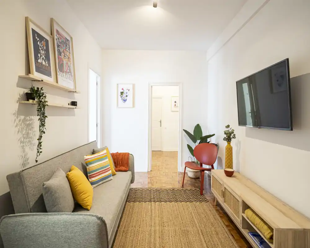 Bright, stylish living room with a comfortable grey sofa, colorful cushions, woven rug, wall-mounted TV and tasteful plants—inviting and well staged for guests.
