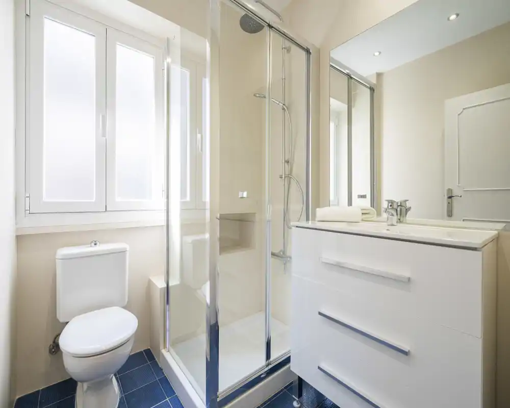 Bright, modern bathroom with a glass-enclosed shower, white vanity with sink and mirror, and a toilet beside a frosted window.