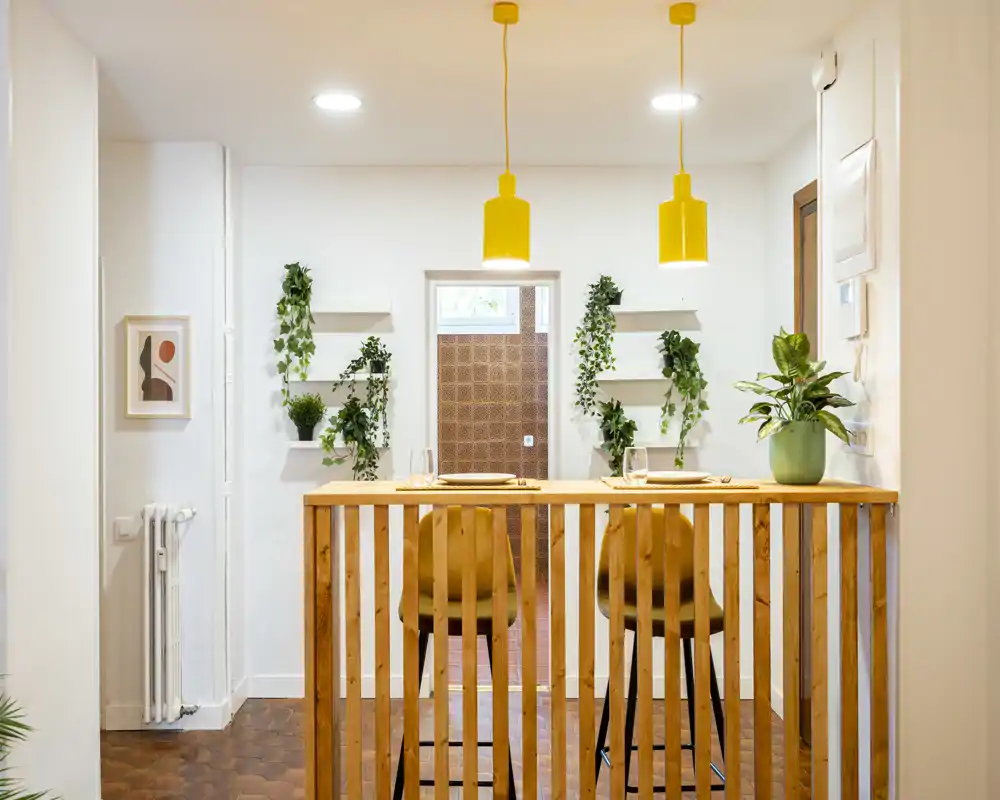 Bright, modern dining area with a wooden breakfast bar, two stools, yellow pendant lights and decorative plants — ideal for casual meals.