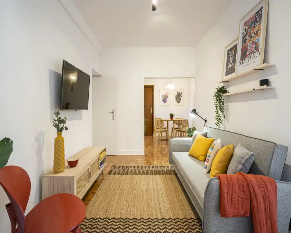 Bright, styled living room with a grey sofa, colorful cushions and a textured rug; wall-mounted TV and a visible dining area beyond, offering a welcoming shared space.
