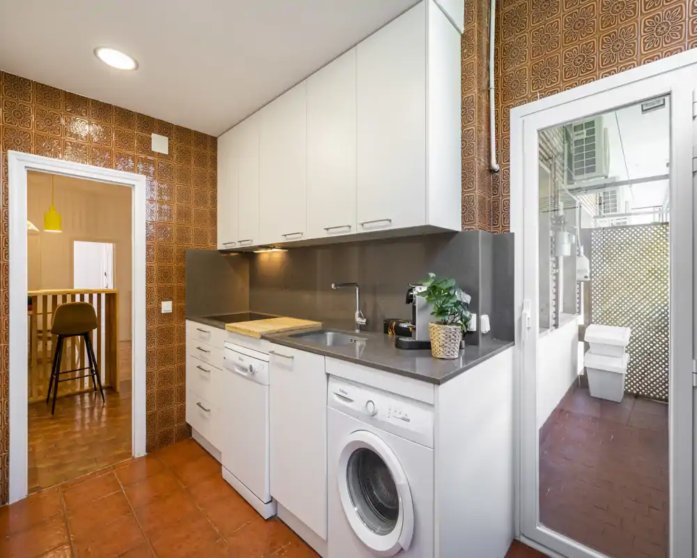 Compact, well-equipped kitchen with white cabinets, integrated sink, washer and dishwasher, warm tiled floor and a door to an enclosed balcony/laundry area.