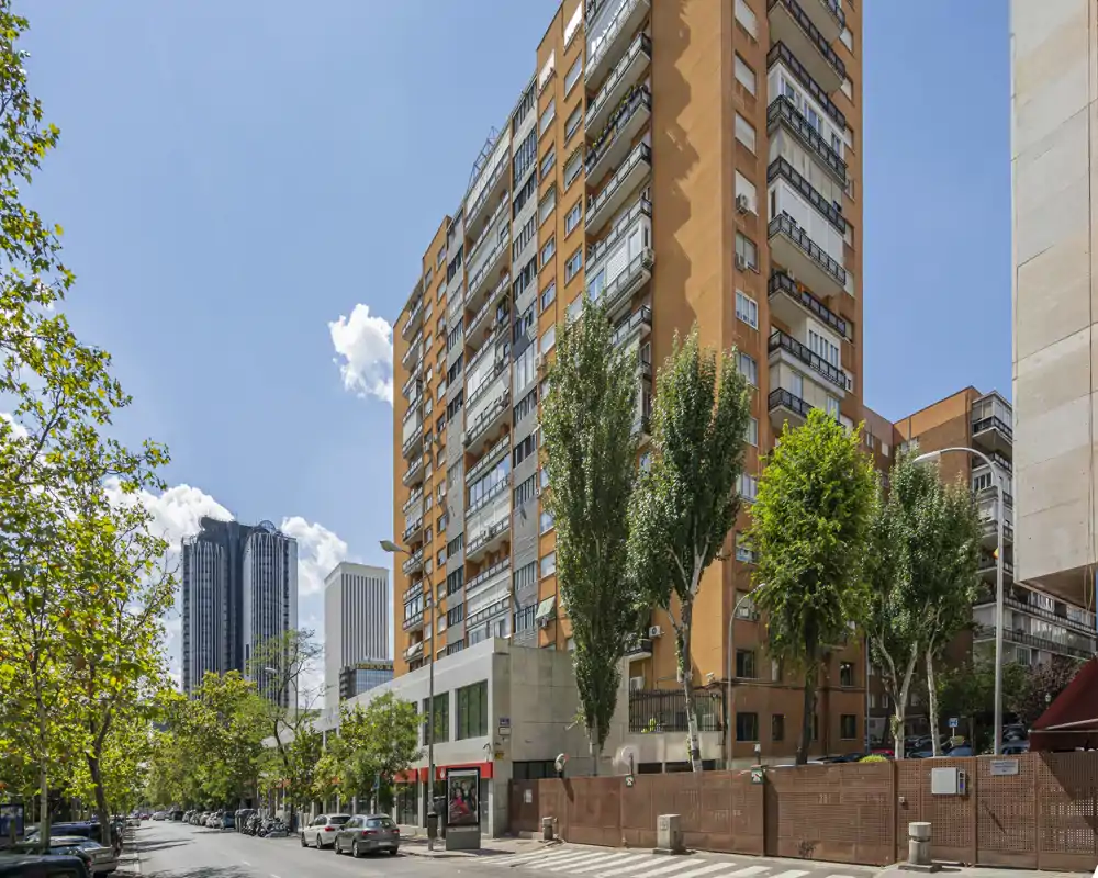 Exterior view of a residential building on a sunny city street, showing balconies, trees and surrounding high-rises.