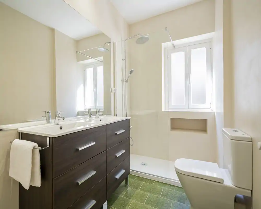 Bright, modern bathroom with a double sink vanity, walk-in shower with glass screen and a window providing natural light.