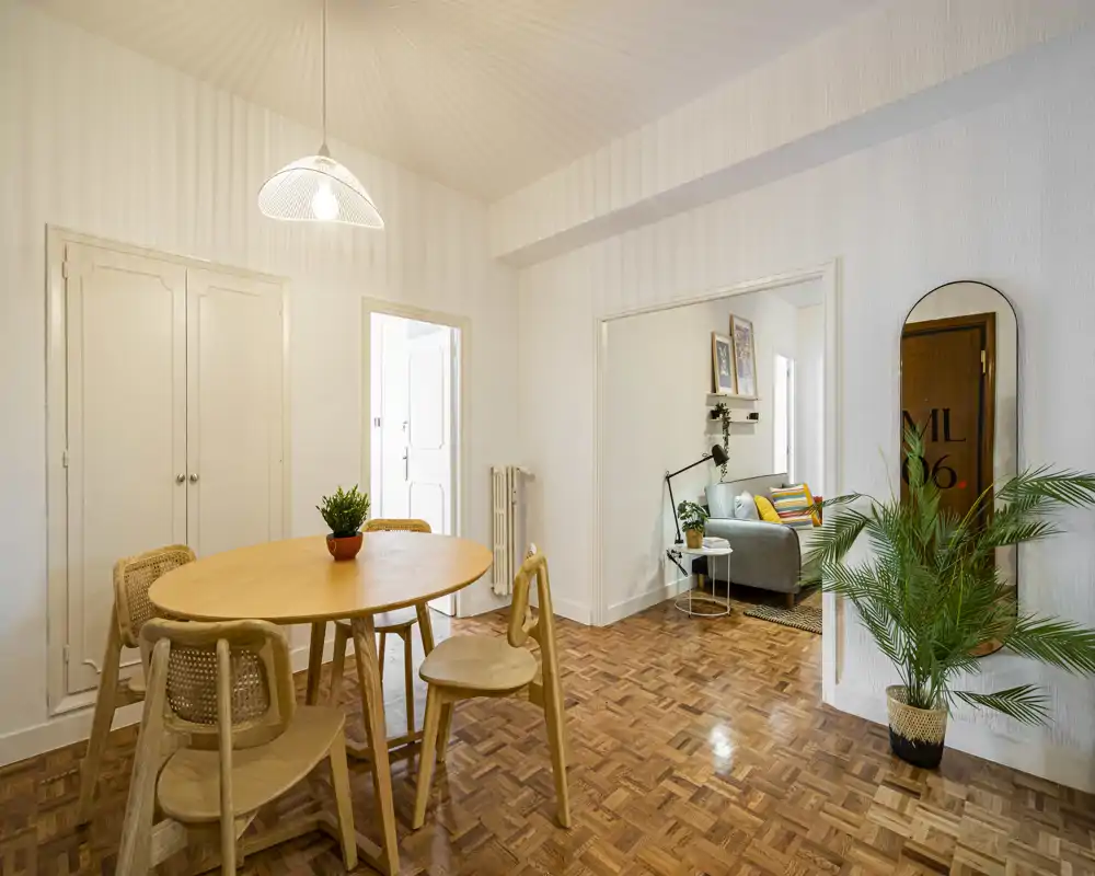 Bright, modern dining area with a round wooden table and woven chairs, warm parquet flooring and greenery creating a cozy, inviting atmosphere.