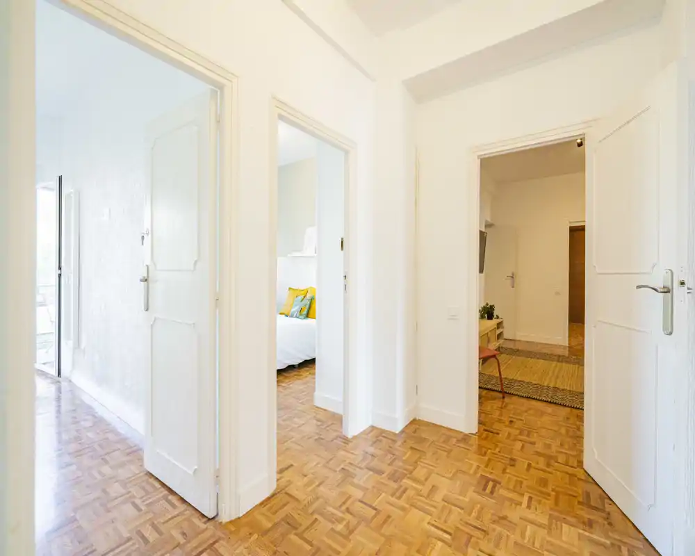 Bright hallway view with open doors revealing a bedroom: you can see a neatly made bed with colorful cushions through the central doorway. Clean parquet floors and white walls create a fresh, inviting feel.