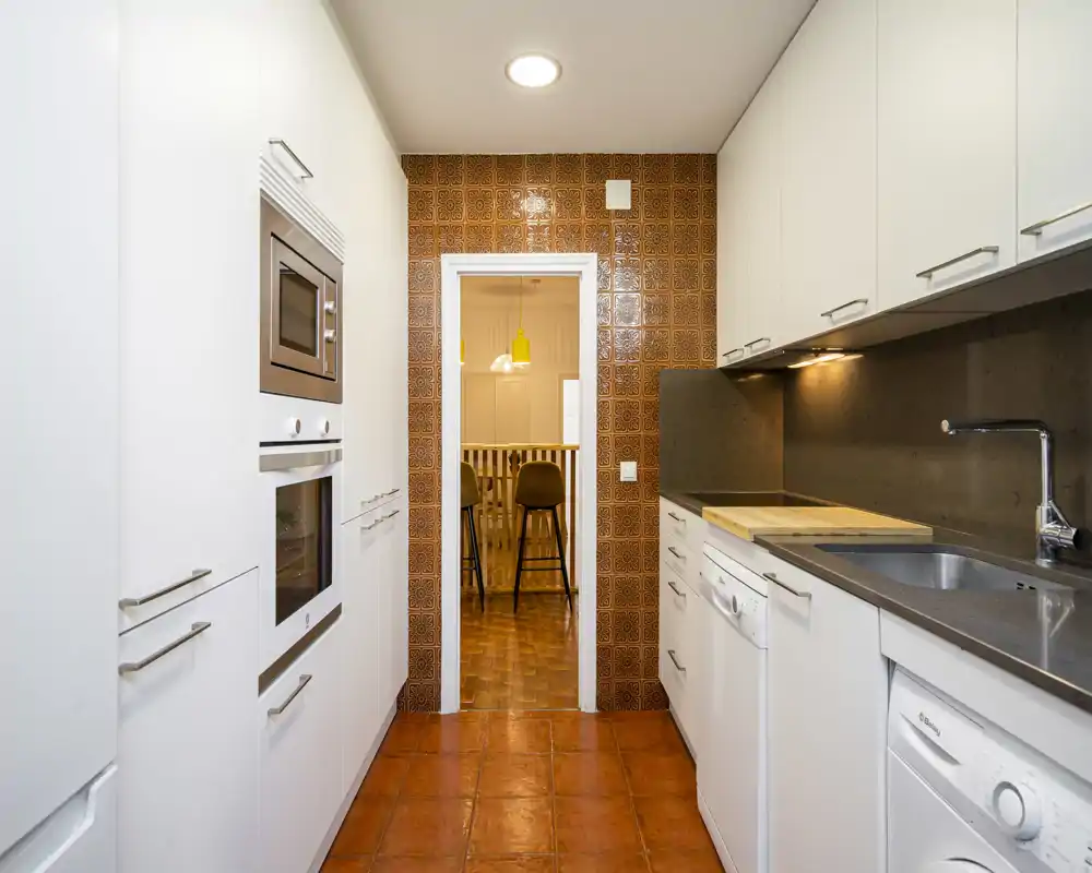 Bright narrow kitchen with white cabinetry, integrated appliances and warm terracotta tiles; practical layout with sink, oven and washing machine included.