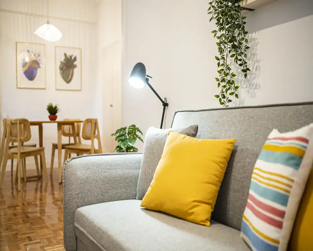 Bright, modern living room with a comfortable grey sofa accented by colorful cushions, a floor lamp and plants; a dining table is visible in the background.