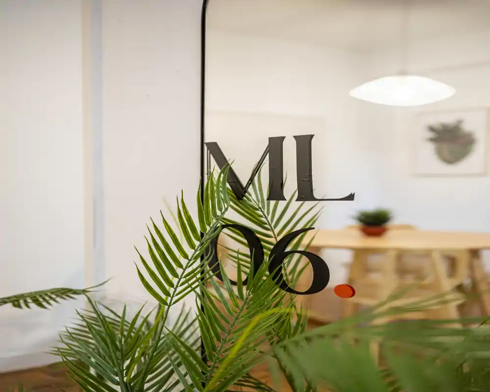 Close-up of indoor greenery against a glass partition with decorative lettering; a blurred dining table and light are visible in the background, creating a calm, styled interior vignette.