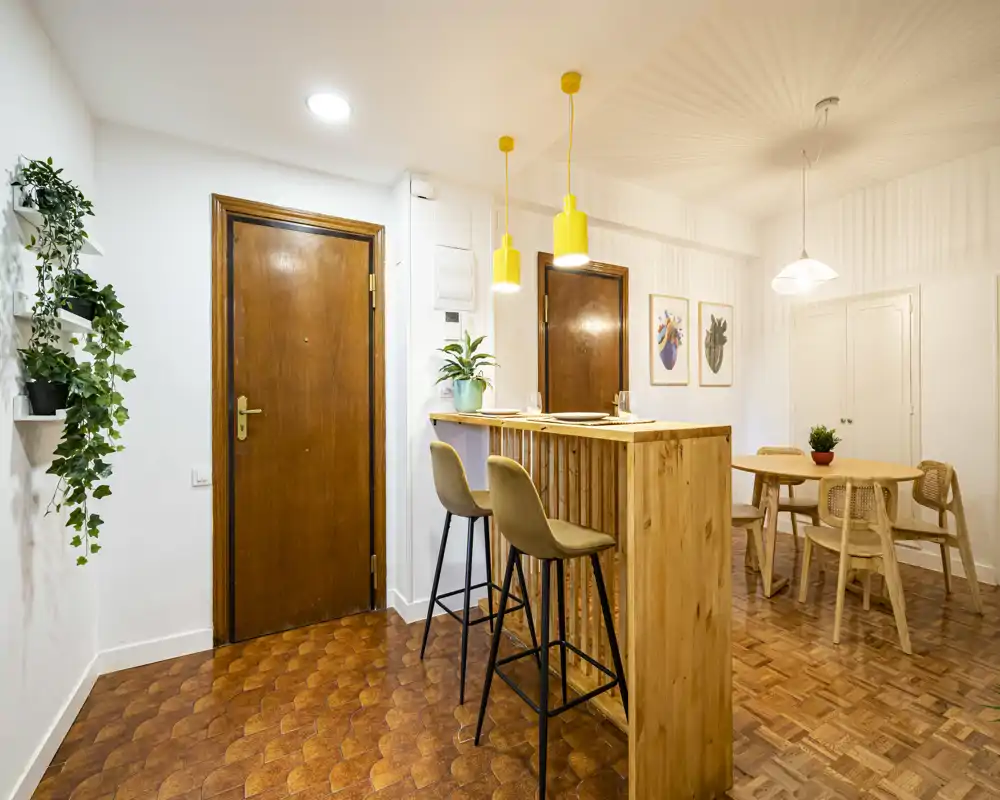 Open-plan dining area with a wooden breakfast bar and high stools, a small dining table with chairs, warm pendant lights and plants creating a cozy, well-lit space.