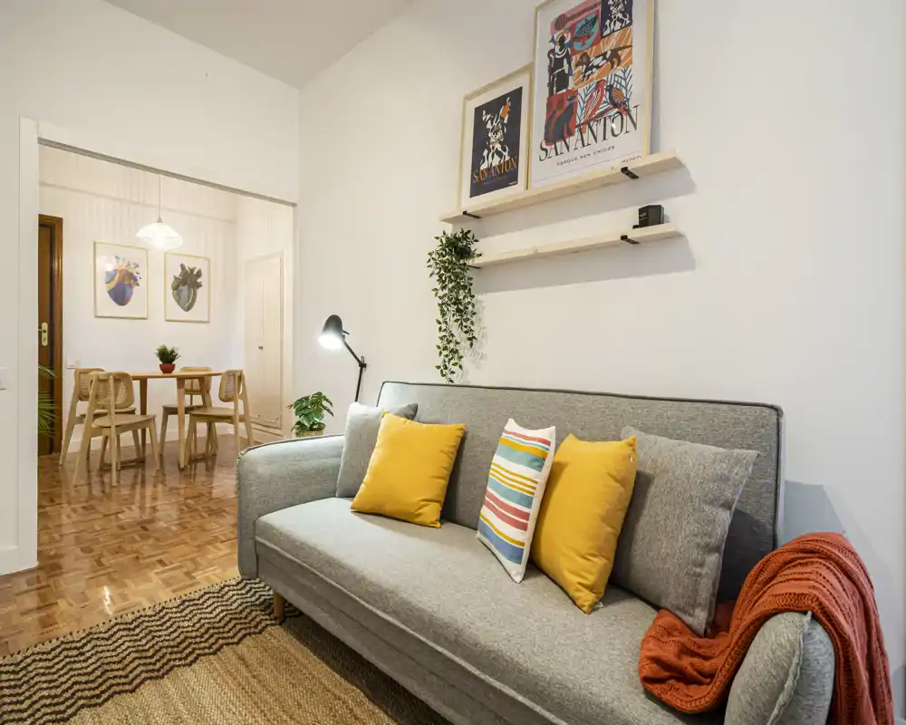 Bright, well-styled living room with a grey sofa, colorful cushions and a warm rug; the adjacent dining area is visible through a wide opening.