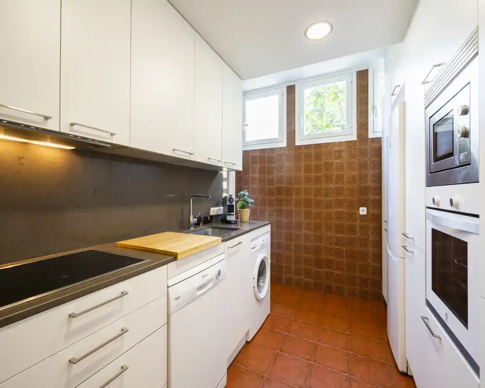 Compact, modern kitchen with white cabinets, integrated appliances, a built-in oven and a washing machine. Terracotta floor and patterned wall tiles give the space a warm, Mediterranean feel.