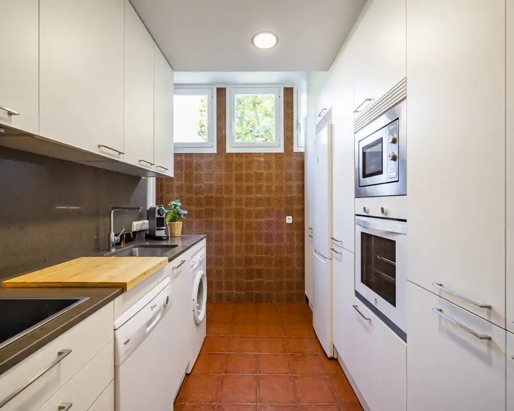 Bright, modern galley kitchen with white cabinetry, integrated appliances and terracotta floor tiles. The layout includes a dishwasher, washing machine, oven and microwave with good natural light from high windows.