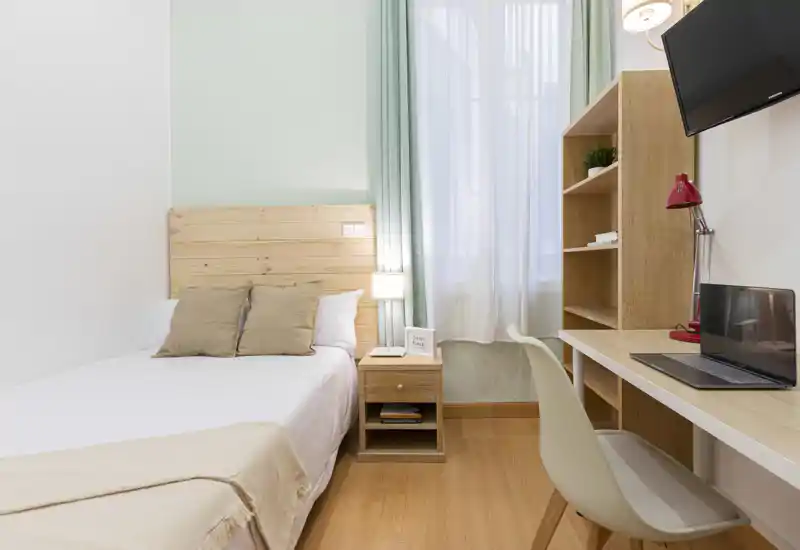 Chambre simple lumineuse et soignée avec tête de lit en bois, table de chevet et un coin bureau pratique près de la fenêtre — parfaite pour un étudiant ou un voyageur seul.