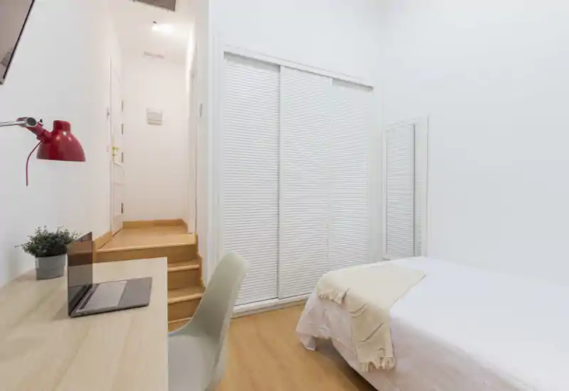 Chambre lumineuse et minimaliste avec un lit simple, une armoire à portes coulissantes et un petit bureau. Le sol en bois et le linge de lit neutre offrent une ambiance calme et accueillante, idéale pour une location courte durée.