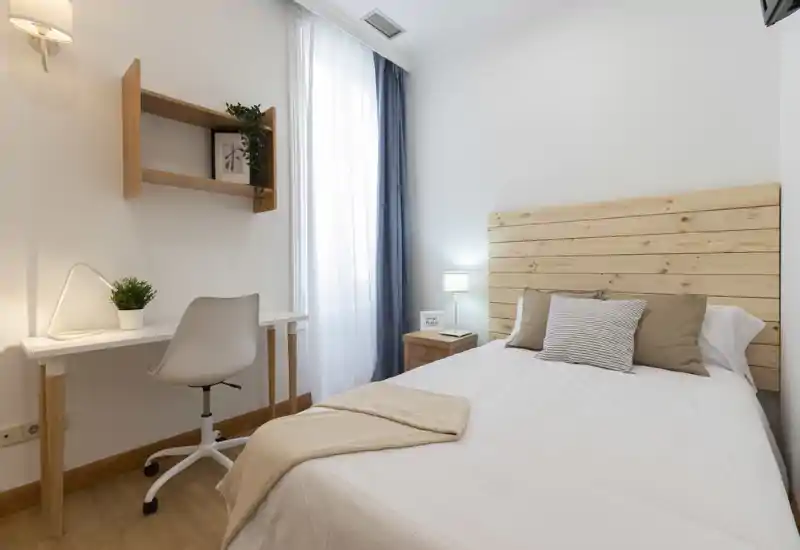 Camera da letto luminosa e ben arredata con testiera in legno, comodino e scrivania. La luce naturale attraverso le tende leggere e i tessuti morbidi creano un'atmosfera accogliente, perfetta per un annuncio.