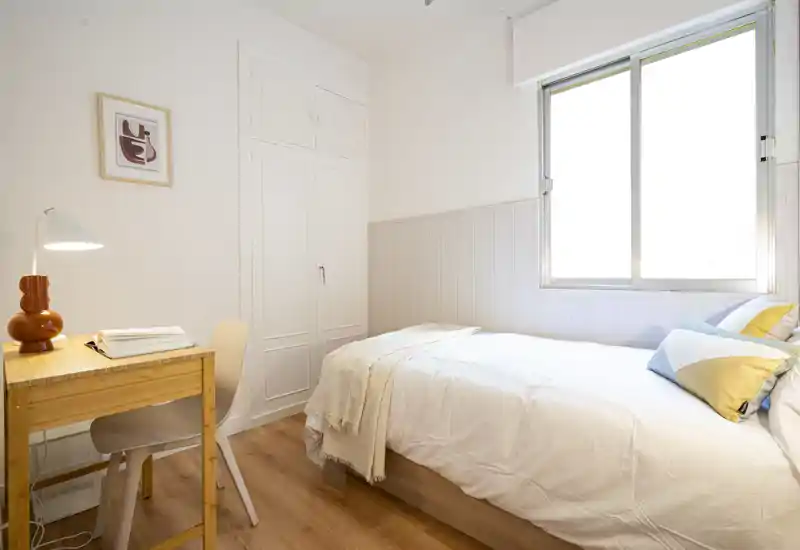 Bright, cozy single bedroom with a neatly made bed, decorative cushions and a small wooden desk by the window — ideal for comfortable short stays.
