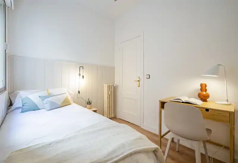 Small, bright bedroom with a neatly made single bed, soft pastel cushions and a wooden desk with lamp — ideal for a cozy, minimalist stay.