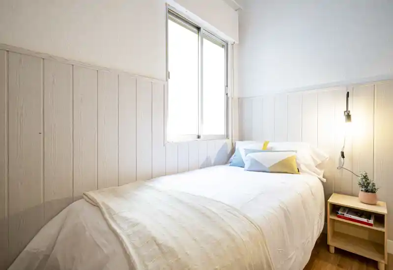 Bright, well-staged bedroom with a neatly made single bed, decorative cushions and a warm bedside lamp providing cozy natural light.