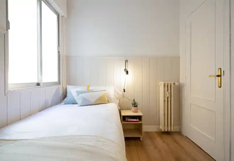 Sunny, well-styled single bedroom with a neatly made bed, bedside table, hanging lamp and large window providing abundant natural light.