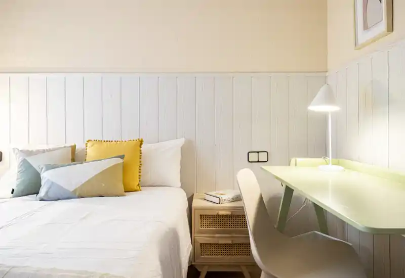 Bright, styled bedroom corner with a neatly made bed, decorative cushions and a small workspace with a lamp — ideal for a cozy single or guest room.