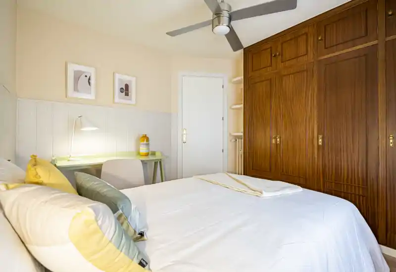 Bright, well-styled bedroom with a large bed dressed in soft linens, decorative cushions and a wooden built-in wardrobe. A small desk with a lamp adds practical workspace while warm tones and natural light create a cozy atmosphere.