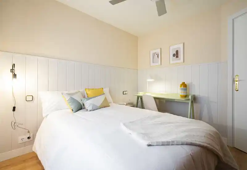 Bright, neatly styled bedroom with a double bed, soft linens and a small work desk. Warm pastel tones and cushions create a welcoming, modern feel.