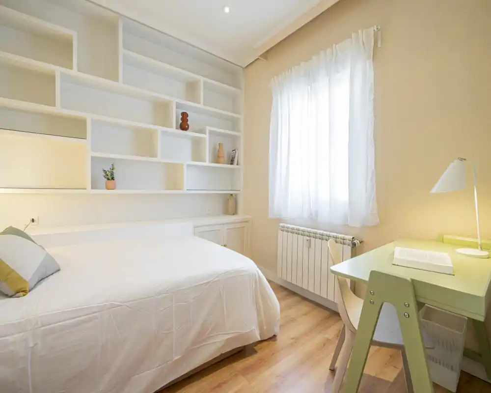 Camera da letto luminosa e minimal con letto matrimoniale, scaffalatura a parete e una scrivania vicino alla finestra — ideale per una persona o uno studente. Pavimento in legno caldo, biancheria bianca e luce naturale che filtra dalla tenda.