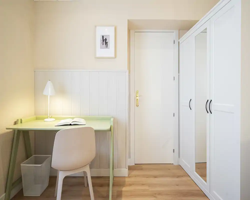 Angolo camera compatto con zona studio e guardaroba: scrivania verde chiaro, sedia, lampada e armadio bianco con specchio creano un'area notte minimal e tranquilla.