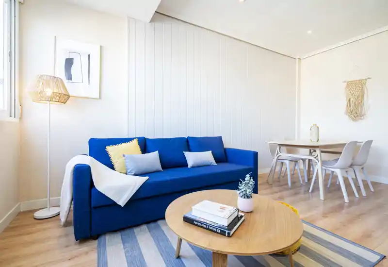 Bright, modern living room with a bold blue sofa, cozy cushions, a round wooden coffee table and a small dining area in the background — ideal for stylish short-term rentals.
