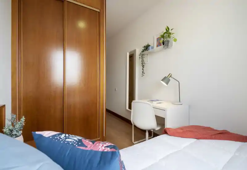 Dormitorio luminoso y ordenado con la cama en primer plano, armario empotrado de madera y un escritorio blanco con lámpara — espacio acogedor y bien presentado ideal para anuncios.