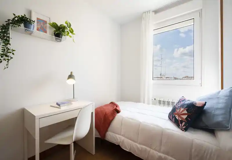 Dormitorio pequeño y luminoso con cama individual bien hecha, cojines decorativos y un escritorio junto a la ventana. Paredes blancas y plantas que crean un ambiente fresco y minimalista, perfecto para una persona.