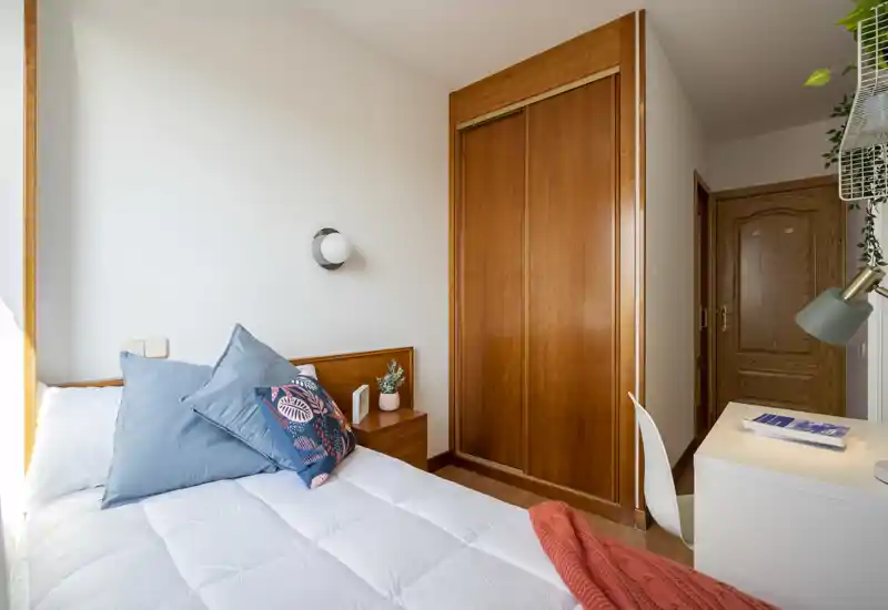 Dormitorio luminoso y compacto con cama bien hecha, armario empotrado de madera y un pequeño escritorio junto a la puerta — acogedor y con buen gusto.