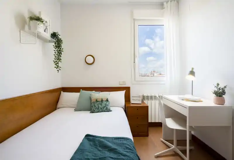 Camera luminosa e compatta con letto singolo con biancheria bianca, cuscini decorativi e plaid verde. Comodino, piccolo scrittoio con lampada e finestra con luce naturale rendono l'ambiente accogliente e funzionale, ideale per affitti brevi.
