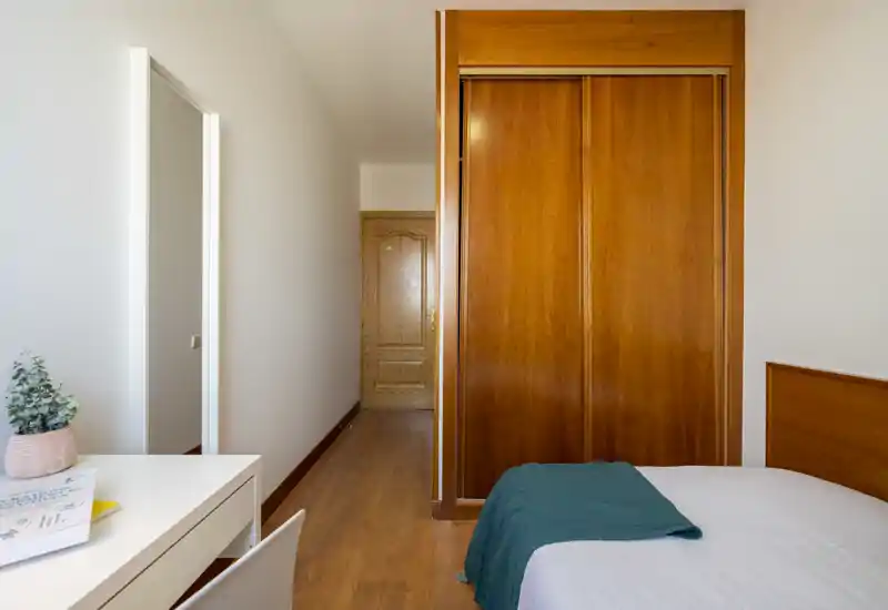 Camera da letto luminosa e minimal con armadio a muro in legno, scrivania bianca e letto rifatto con una coperta verde petrolio. La luce naturale e il pavimento in legno creano un ambiente accogliente e ordinato, adatto per annunci.