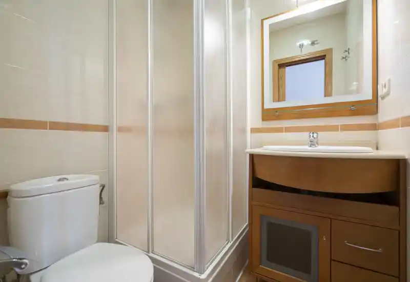 Salle de bain compacte et propre avec douche d'angle, WC et meuble vasque en bois avec miroir — lumineuse et fonctionnelle pour de courts séjours.